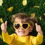 Ein kleiner Junge im Kindergartenalter liegt entspannt auf dem Rasen und streckt mit einem Lächeln im Gesicht und einer Sonnenbrille auf der Nase beide Daumen in die Luft.