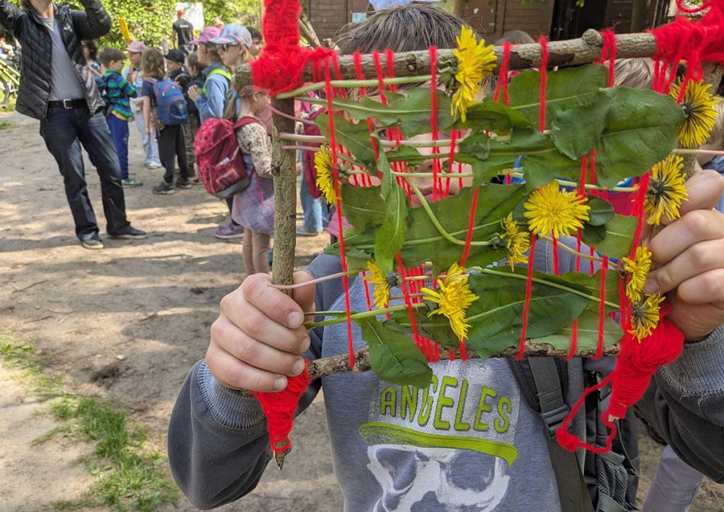 Ein Junge im Schulalter hält einen Webrahmen aus Naturmaterialien vor sein Gesicht und präsentiert sein Arbeitsergebnis.