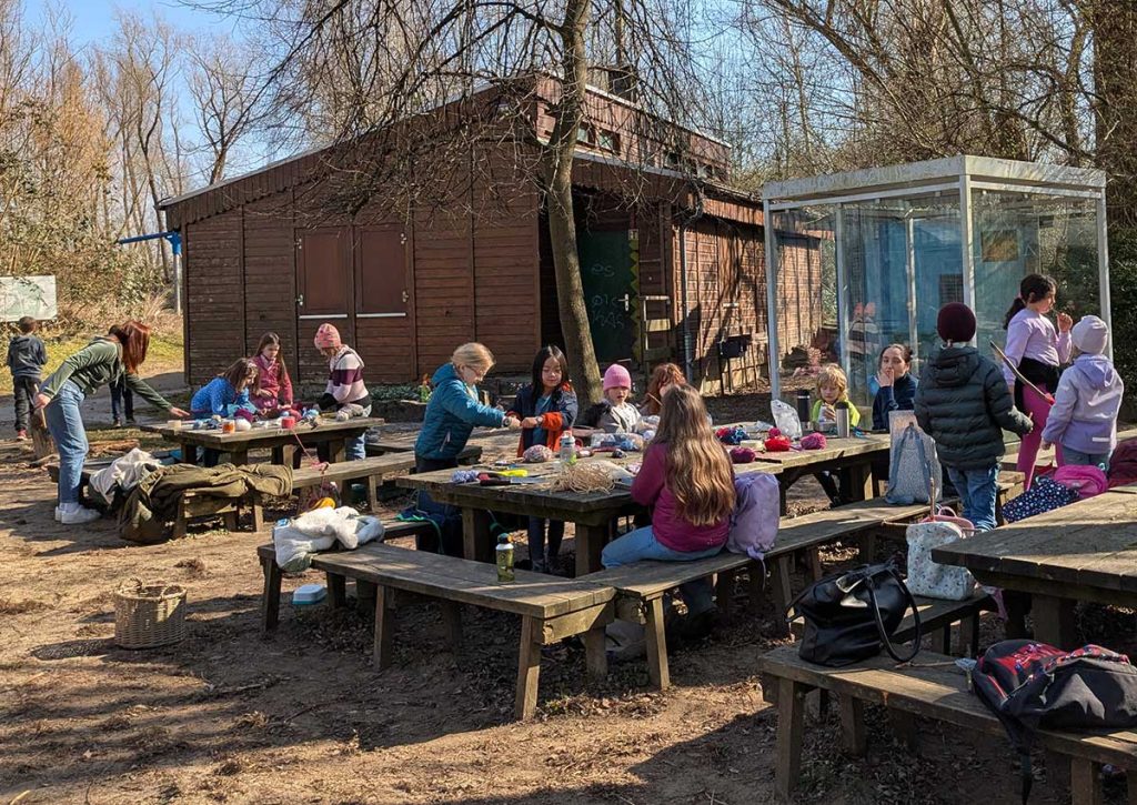 Schulkinder sitzen auf einem Abenteuerspielplatz draußen an großen Tischen und stellen Webrahmen aus Naturmaterialien her.