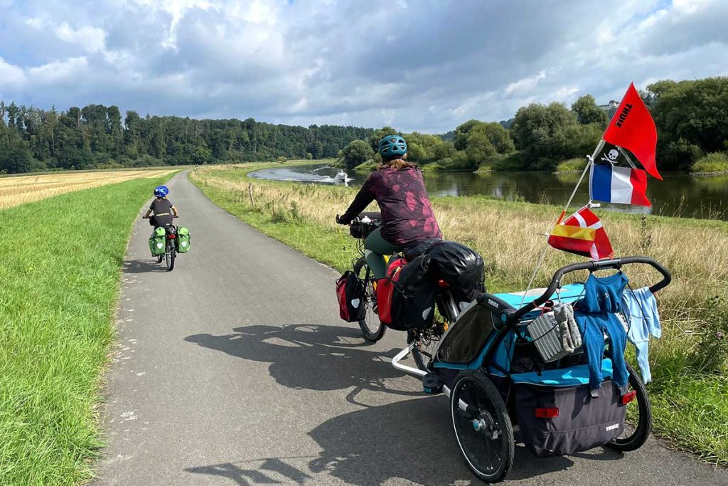 Mutter und Kind fahren mit Gepäck und Radanhänger in idyllischer Landschaft entlang an einem Fluss.
