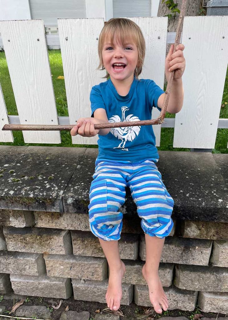 Ein Junge im Grundschulalter schaut fröhlich in die Kamera und hält dabei begeistert einen Stock in der Hand mit dem er spielt.