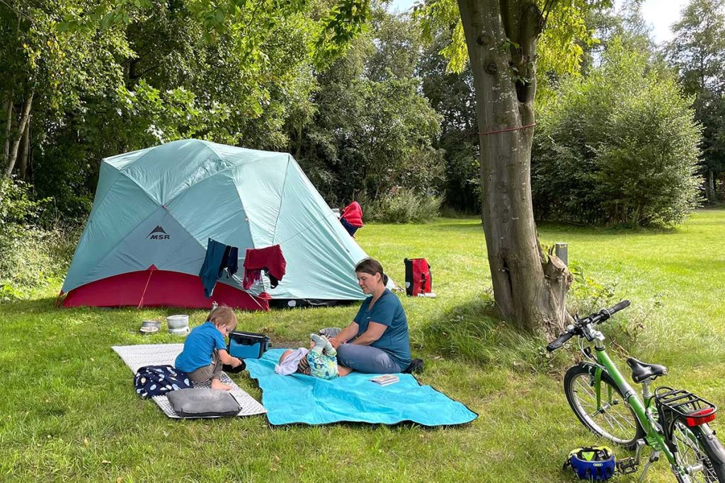 Ein Zelt auf einer Wiese in gemütlicher Atmosphäre sitzen Mutter und Kleinkind auf einer Decke vor dem Zelt und picknicken. Rechts von ihnen ist ein Kinderfahrrad zu sehen.
