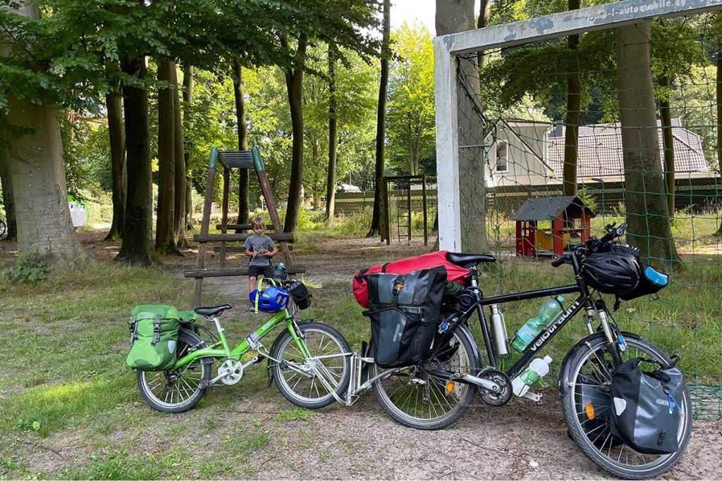 Zwei bepackte Fahrräder - eines davon ein Kinderfahrrad - stehen auf einem Spielplatz an ein Tor angelehnt. Im Hintergrund ist ein Spielplatz zu sehen.