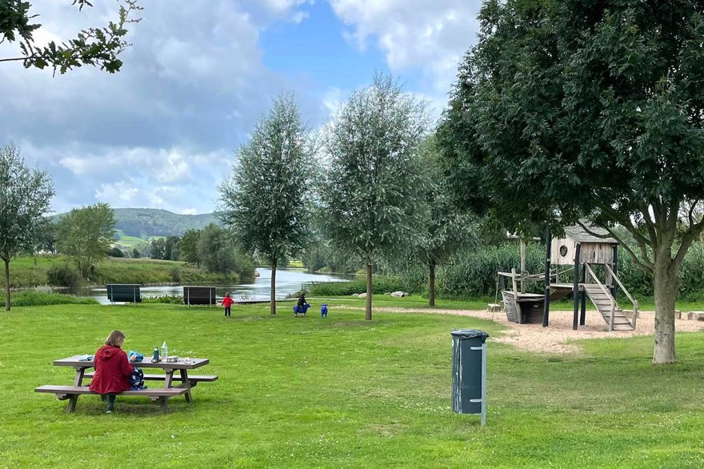 Kleiner Spielplatz mit grüner Wiese, idyllisch gelegen an einem Fluss. Die Mutter sitzt an einem Picknicktisch und pausiert. Ein Kind befindet sich auf der Wiese.