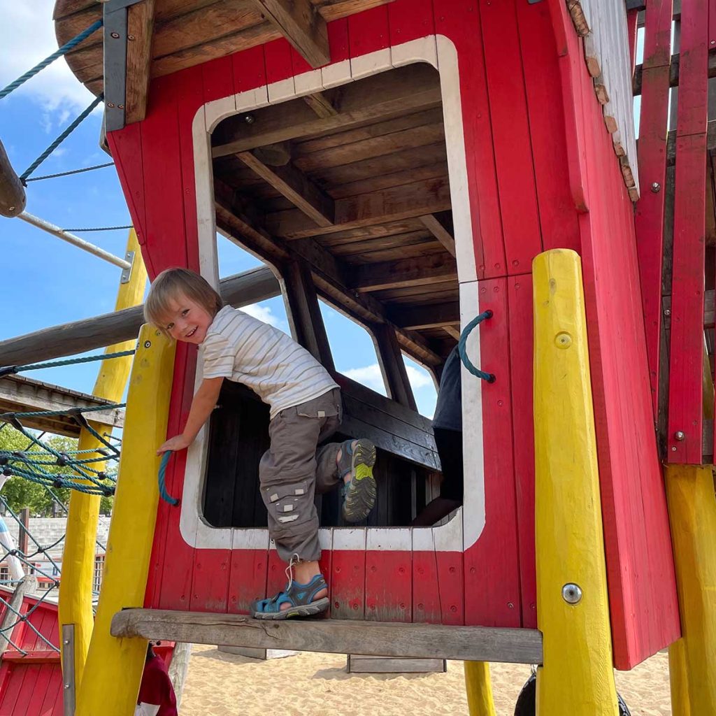Ein Junge im Grundschulalter klettert fröhlich auf einem Holzklettergerät auf dem Spielplatz Vegesacker Krabbe in Bremen.