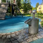 Playground with a wooden climbing structure and a water fountain spraying water into a gray basin on a blue safety surface.