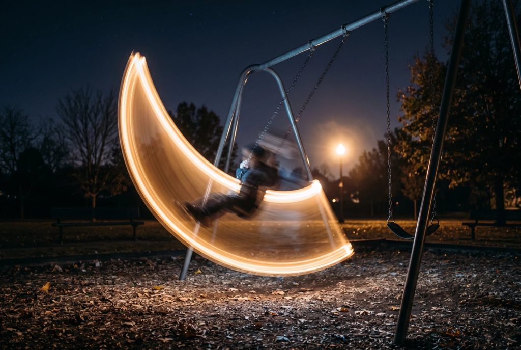Langzeitbelichtung an einer Schaukel bei Nacht: Eine Lampe zieht einen leuchtenden orangefarbenen Bogen, die schaukelnde Person ist nur verwischt zu sehen.