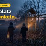 Zwei Kinder in Winterjacken stehen von hinten gesehen mit Taschenlampen und einer Laterne vor einem Holz-Spielturm in der Abenddämmerung.